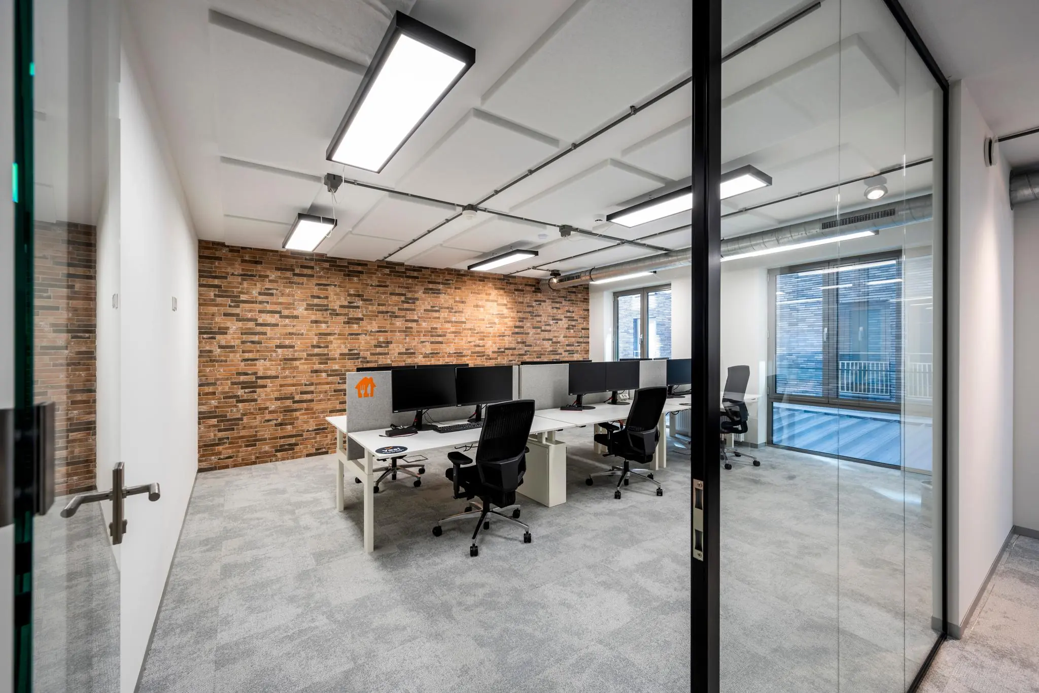 Modern kantoor met werkplekken, bureaustoelen en computerschermen aan de Harry Banninkstraat, met een bakstenen accentmuur en glazen wanden.